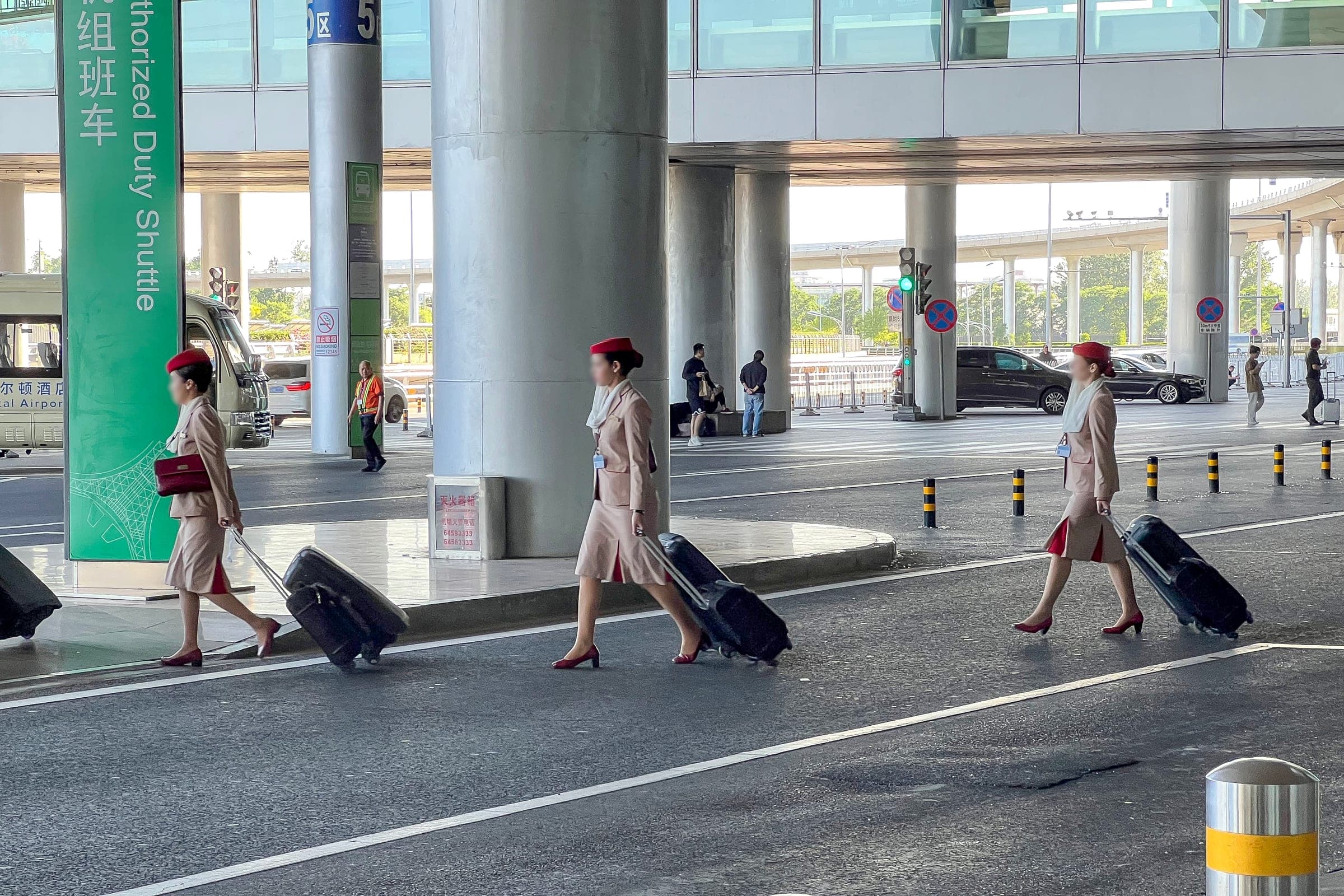 Three Emirates cabin crew in beige uniforms and red hats walk in single file at Beijing Capital Airport's Terminal 3, each pulling a wheeled suitcase towards the crew bus.