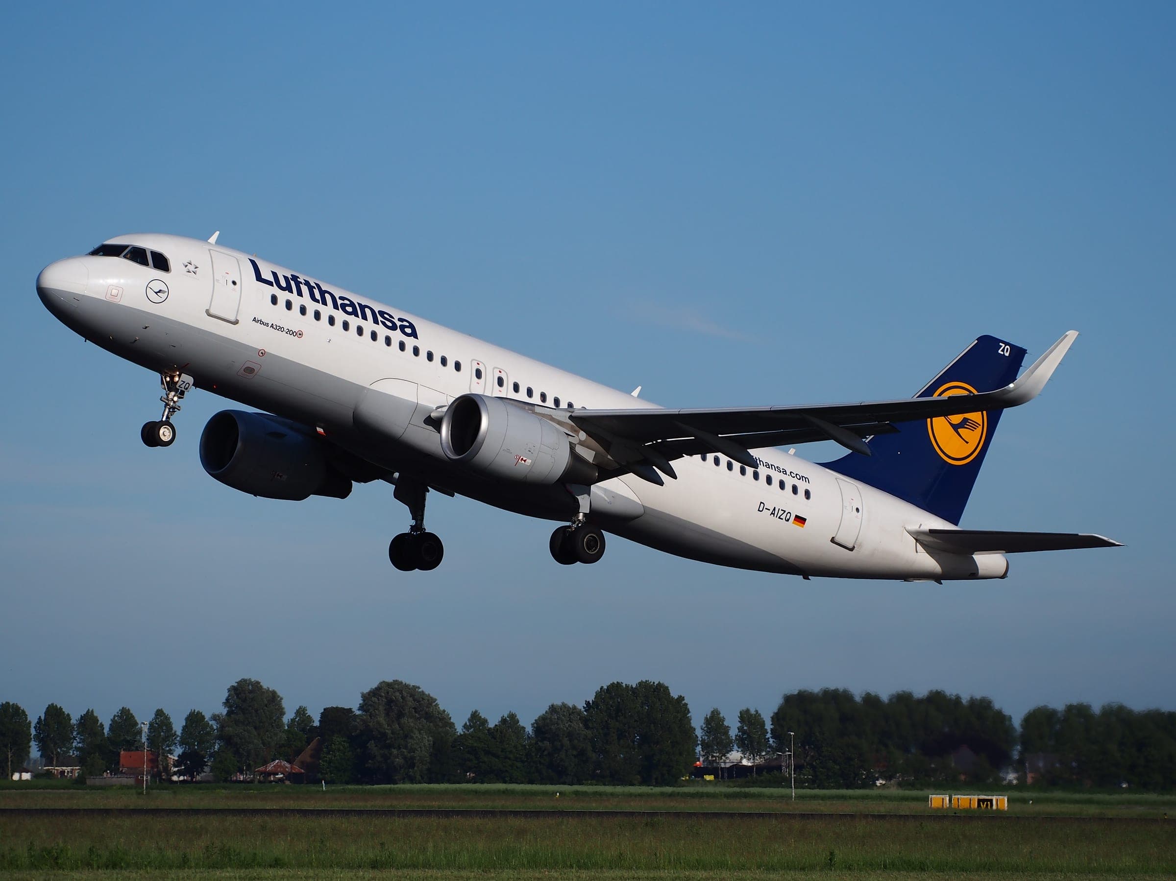 A Lufthansa Airbus A320 rotating off the runway at Amsterdam Schiphol on a clear evening, treeline behind.