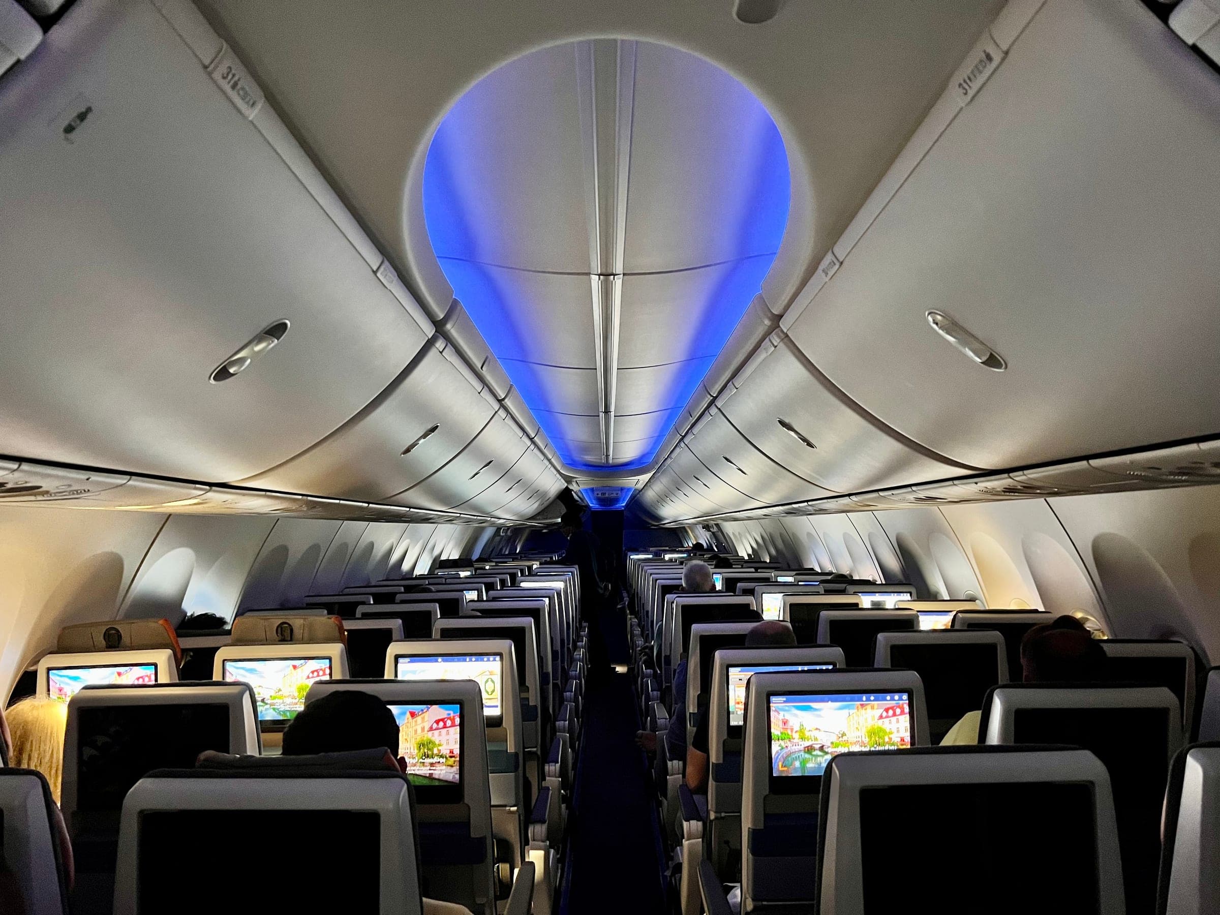 The economy cabin of a flydubai Boeing 737 MAX seen from a seat near the back, looking forward along the aisle beneath blue-lit overhead lockers, with rows of lit seatback screens and a few passengers seen from behind.