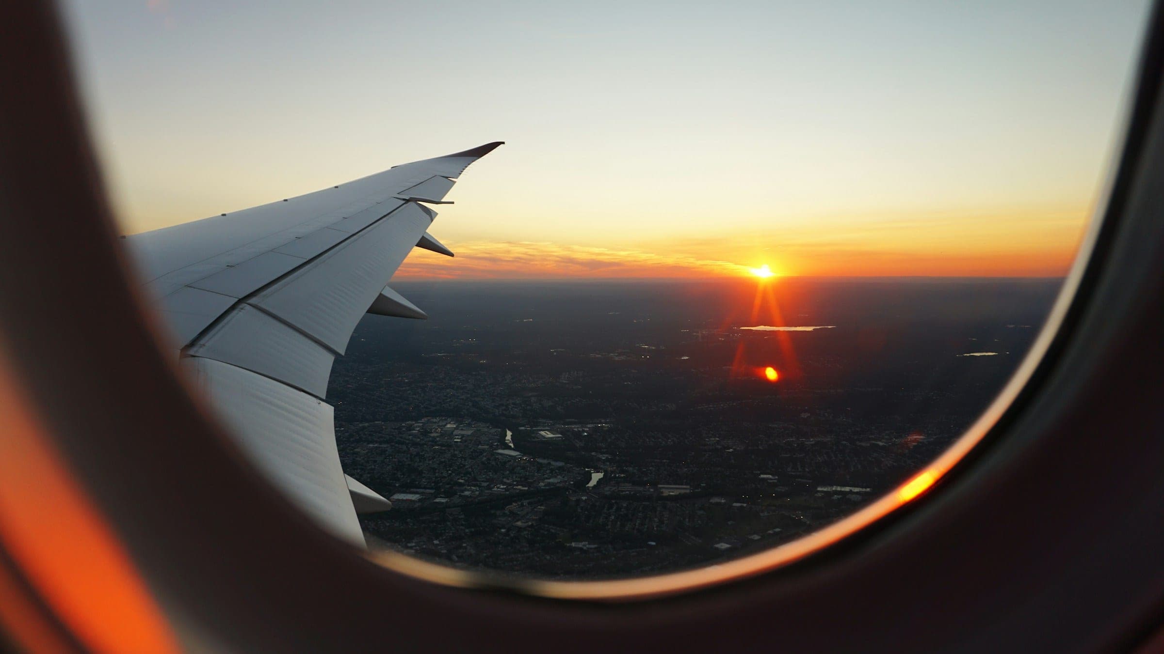 An airliner's wing and a low sun seen through a cabin window, the land below already in shadow at dusk.